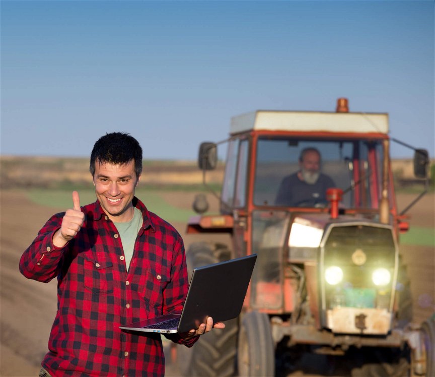 Junger Bauer steht mit Laptop vor einem Traktor und streckt den Daumen freudig nach oben