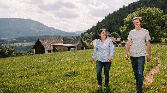 Der Ursprungsbauer Ronald und die Murauer Ursprungsbäuerin Claudia Sperl spatzieren über eine gründe Wiese hinter einem Bauernhof